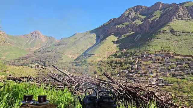 Small tea cups rest on a flat stone slab beside a blazing campfire with kettles, overlooking a terraced village and rugged mountain peaks under a clear blue sky.
📍 Kamalla village, Hawraman Takht