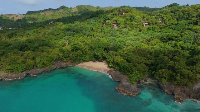 Crescent beach nestles between rocky headlands at Playa del Amor in Las Galeras Samana Dominican Republic, hillside villas peek through dense canopy above turquoise cove. Aerial drone view.