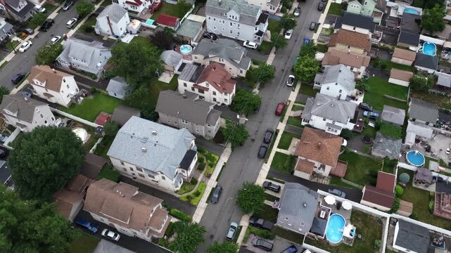 Suburban street winds past red house and backyard pools in Belleville New Jersey, cars travel pavement between homes with rooftop solar panels and gabled roofs below. Aerial drone top down flyover