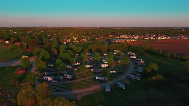 Aerial drone view at sunset over an RV park, fifth wheels and travel trailers along circular gravel loops in golden hour light, with a small Midwest town and farmland beyond.