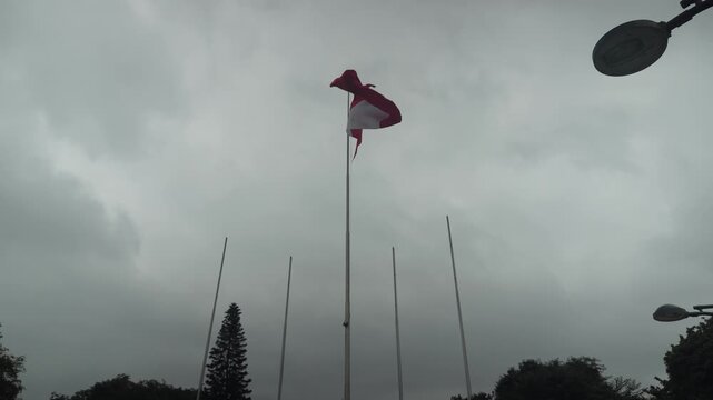 A big red and white Peruvian flag waving in the wind in a public park. 4 other shorter flag poles stand next to the centered taller one. Recorded on a cloudy morning in Lima, Peru.