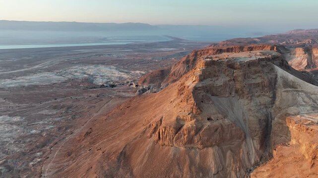 Drone camera pans slowly past high altitude rock crags revealing a vast desolate valley and distant dead sea shoreline under a soft morning horizon sky.