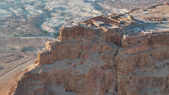 High angle drone shot focusing on the sheer vertical limestone cliffs and ancient archaeological complex of Masada overviewing the rugged terrain below at morning golden hour.
