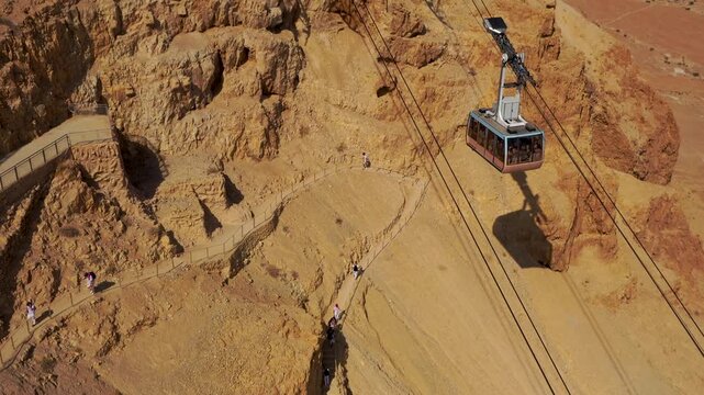 Drone shot starting top down on tourists climbing the snake path before tilting up to track a moving cable car cabin ascending the rugged desert cliffs toward the upper terminal station