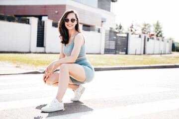 Fototapeta premium Young beautiful smiling female in trendy summer blue cycling shorts and tank top clothes. Carefree woman posing in street. Positive model having fun. Cheerful and happy. Sits on asphalt. In sunglasses