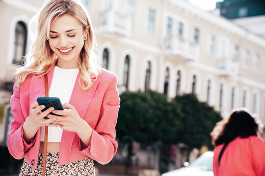 Young beautiful smiling hipster woman in trendy summer jacket clothes. Carefree model posing in street at sunset. Positive blond female. Holds smartphone, uses phone apps, looks at telephone screen