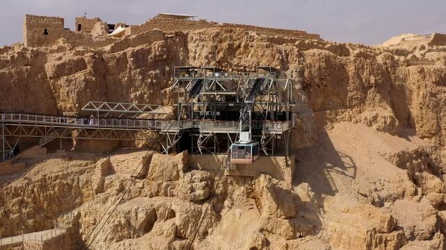 High angle drone shot capturing the modern cable car station built into the rugged cliffs of Masada fortress with a passenger cabin arriving at the upper terminal