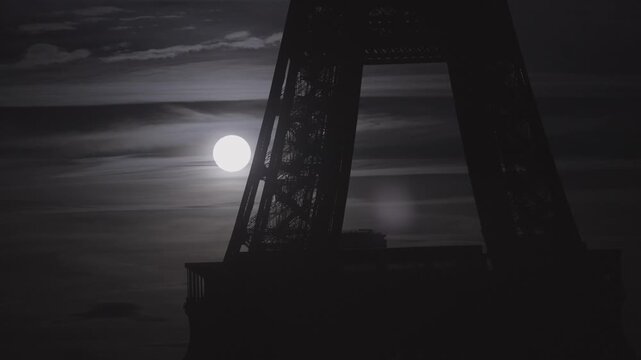 The full moon glows through the iron latticework at the base of the Eiffel Tower at dusk, creating a striking silhouette.
