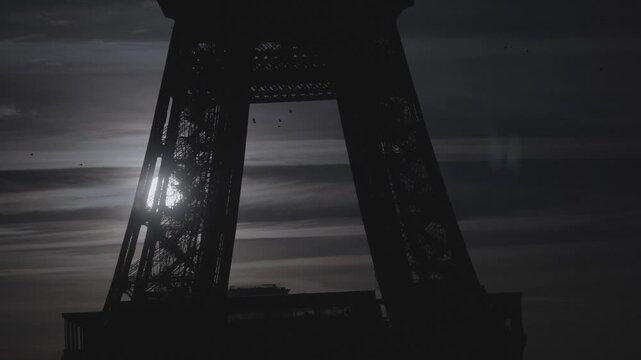Soft sun flare filters through the intricate iron lattice at the base of the Eiffel Tower against a hazy dusk sky.