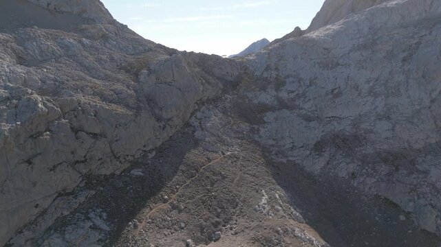Aerial drone pull back shot reveals the majestic Picos de Europa mountains in Asturias, Spain. Rugged, rocky terrain around Diente de Urriellu under a bright sky, with a remote hiking trail visible.