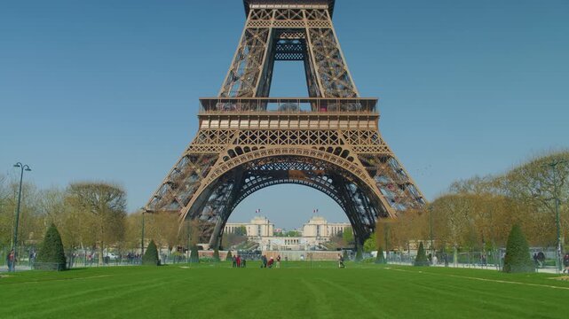Eiffel tower base green lawn people walking background, handheld shot. Scenic view of Parisian landmark steel structure with tourists on grass under daylight.