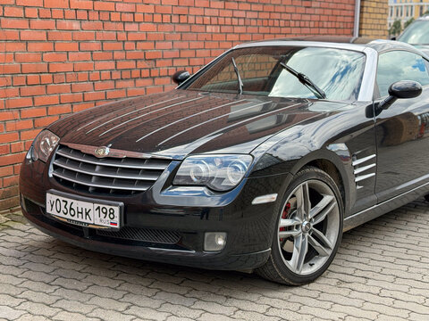 A black Chrysler Crossfire sports car parked on a paved lot near a red brick wall. The vehicle shows its classic coupe design lines, silver alloy wheels, and distinctive front grille under daylight.