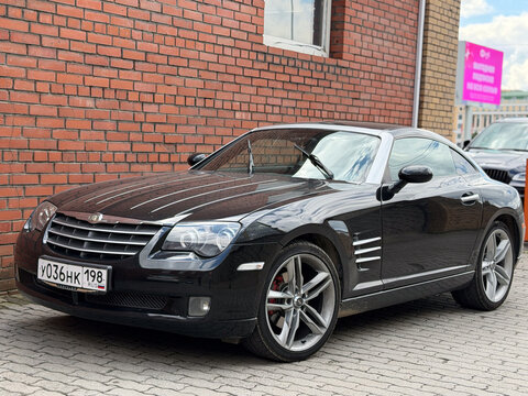 A black Chrysler Crossfire sports car parked on a paved lot near a red brick wall. The vehicle shows its classic coupe design lines, silver alloy wheels, and distinctive front grille under daylight.