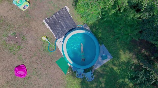 High angle top-down aerial static shot captures a pre-teen girl swimming and sitting inside the clear blue water of a round inflatable pool on a sunny green lawn in a domestic backyard garden