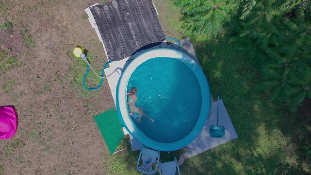 High angle top-down aerial camera movement zooms from a wide yard view to a close-up of a pre-teen girl swimming in the clear blue water of a round inflatable pool in a sunny green backyard garden.