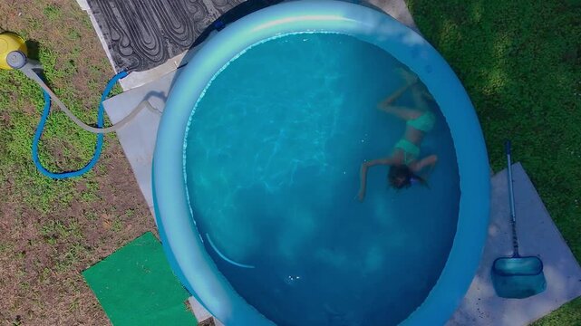 High angle top-down aerial close up shot captures a pre-teen girl swimming underwater, practicing healthy active breaststroke movements inside the clear blue water of a round inflatable backyard pool.