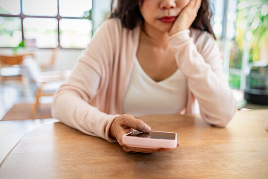 Cropped shot of a bored young Asian woman resting her chin on hand, holding a smartphone while waiting for a message