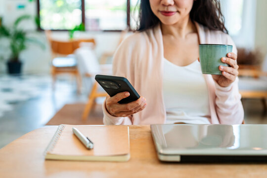 Young woman holding a coffee cup and checking her smartphone at a desk with a laptop and notebook; digital nomad and remote working concept