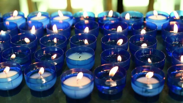 Rows of lit votive candles in blue glass holders inside a church. Slow camera movement captures glowing flames in a quiet religious setting.