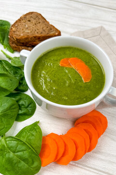 Healthy spinach soup served with bread and carrot slices on a white wooden table