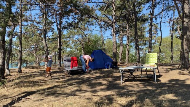 A mother and child play around their blue tent at a campsite. The young boy runs joyfully with a racquet in a sunny pine forest. The active outdoor setting feels happy and peaceful.