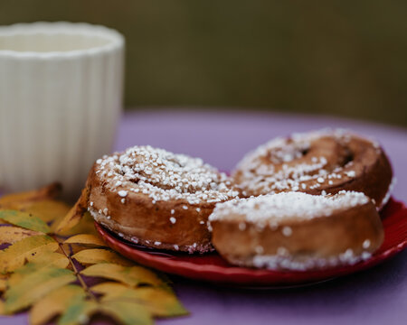 Fresh Swedish cinnamon buns with pearl sugar served on a red plate beside a white coffee mug. Cozy autumn fika scene with colorful leaves and shallow depth of field.