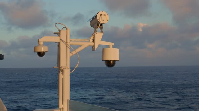 Surveillance cameras mounted on a cruise liner mast overlook open ocean at dusk. White PTZ dome cameras and spotlight attached to metal pole sway gently against dramatic cloudy sky and deep blue sea.