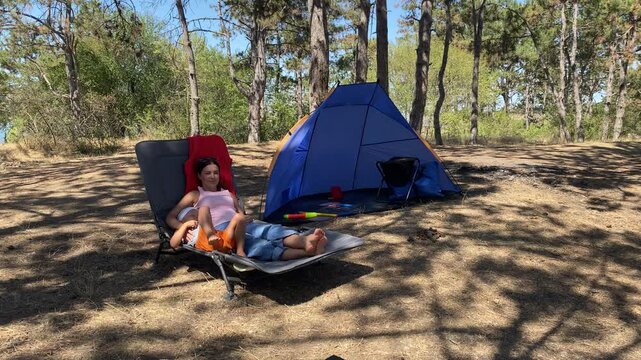 A mother and her child lie together on a lounge chair to rest near a blue tent. They share a cozy, peaceful cuddle in a sunny pine forest. The atmosphere is warm and serene.