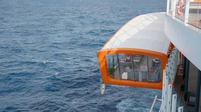 Modern luxury cruise ship features striking orange-framed glass observation lounge cantilevered over calm deep blue ocean waters, with comfortable seating visible inside and white dome canopy above.
