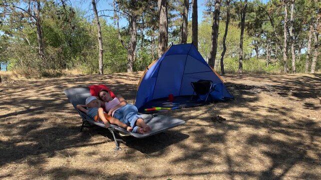 A serene mother and her young boy lie comfortably on a lounger cot. They enjoy a peaceful moment together beside a blue tent. Tall pine trees shade the warm forest campsite.