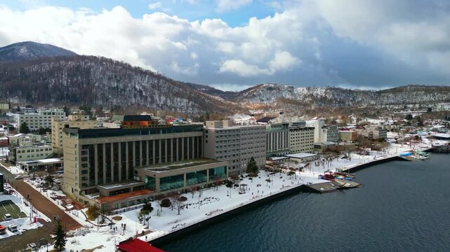 High angle drone shot tracking along the snow-covered promenade, docks, and large lakeside resort hotels with mountains in the background.
