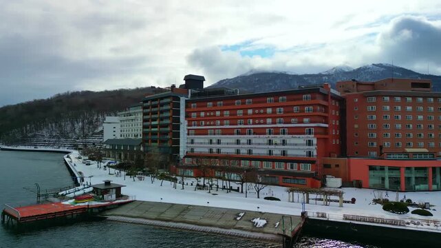 Aerial pan of a modern red lakefront resort hotel complex nestled along a snow-covered shoreline with mountains behind.