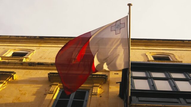 A Maltese flag waves above a sunlit historic facade in warm golden hour light.