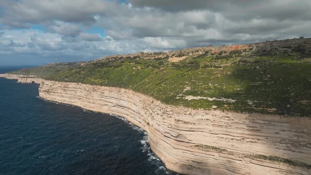 Aerial coastline with layered white cliffs, green slopes, and dark blue sea beneath cloudy skies. Dingli cliffs