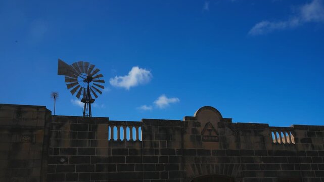 A static stone building topped with a windmill stands under a bright blue sky with scattered white clouds, showcasing rustic architecture and peaceful weather.