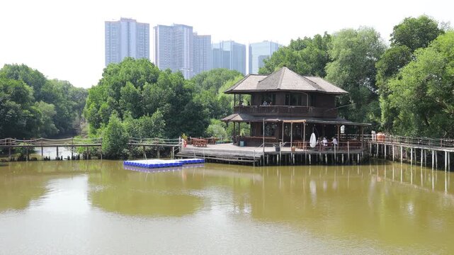 Traditional two-story wooden villa and boardwalk surrounded by lush mangrove forest with modern city skyscrapers in the background in Jakarta.
