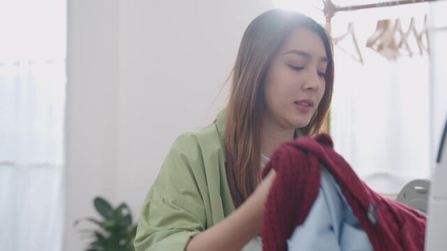 Happy Asian woman holding and smelling freshly washed jean sweater with eyes closed, smile and satisfaction after unloading from washing machine in bright cozy laundry room at home