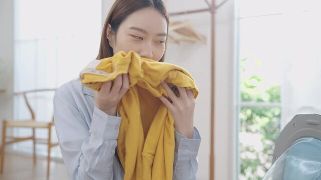 Young happy Asian Japanese woman holding and smelling freshly washed yellow sweater with eyes closed, smile and satisfaction after unloading from washing machine or dryer in cozy laundry room at home