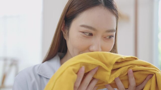 Young happy Asian Japanese woman holding and smelling freshly washed yellow sweater with eyes closed, smile and satisfaction after unloading from washing machine or dryer in cozy laundry room at home