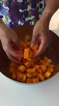 Hands mixing seasoned butternut squash cubes inside a wooden bowl during recipe prep.