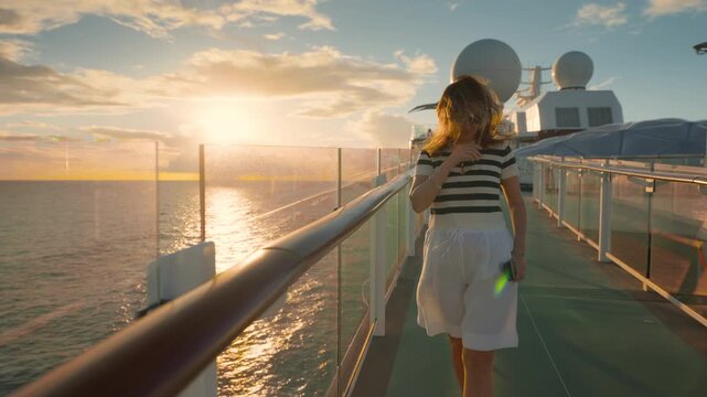 Young woman in striped shirt walks along cruise liner deck at golden hour in Caribbean Sea. Wind blows through her hair as she admires ocean sunset view with glass railing and satellite domes visible.