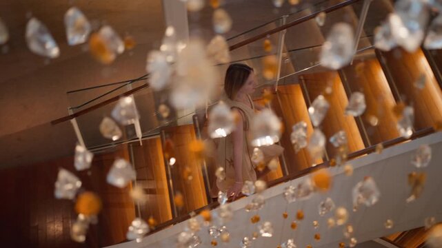 Elegant woman in flowing beige dress descends a curved wooden staircase with glass railings in a luxury hotel lobby, filmed through sparkling crystal and amber chandelier ornaments in foreground.