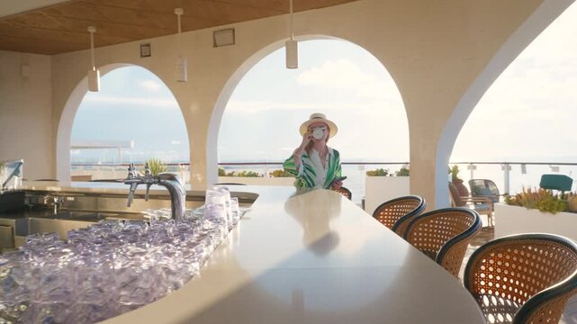 Stylish woman wearing straw hat and tropical print blouse relaxes at Mediterranean-style outdoor bar with white arched columns and green tile decor, sipping coffee while browsing phone on sunny day.