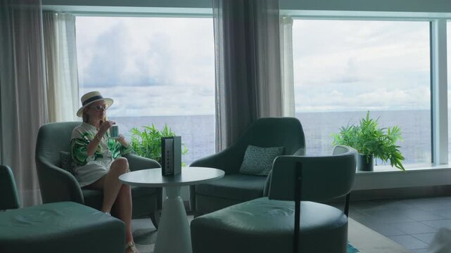 Stylish woman wearing straw hat and tropical print cover-up relaxes in plush teal armchair sipping beverage in elegant cruise ship lounge with panoramic ocean views and lush green plants by windows.