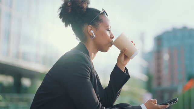 Woman listening to voice message by earphones and smartphone in city street. Office worker or businesswoman relaxing after long working day, sitting in park and drinking coffee or tea for recovery