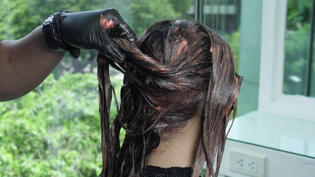 Hair coloring treatment in progress as mahogany brown or dark auburn dye is applied to a Southeast Asian woman&rsquo;s hair at a professional salon.