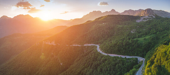 Naklejka premium Winding mountain road traversing a lush green forest with dramatic sun flare and mountain peaks during a vibrant sunset