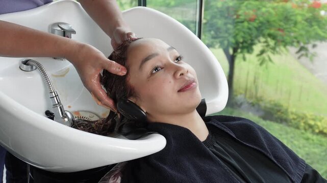 Hair stylist massaging mahogany brown dyed hair before rinsing during professional hair coloring treatment for Southeast Asian woman in her early 20s