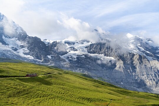 Jungfrau Railway to Jungfraujoch runs through green meadows, above the Kleine Scheidegg stop, rack railway, in the back mountains with peaks Eiger, M&ouml;nch and Jungfrau, Jungfrau region, Bernese Oberland, Switzerland