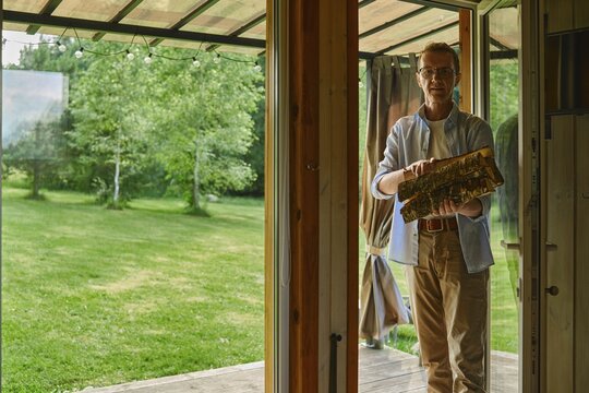 Middle-aged man holds stack of firewood while standing at entrance of wooden cabin. He is preparing wood for fireplace. Green grass and trees fill backyard area. Clear skies show sunlight coming through